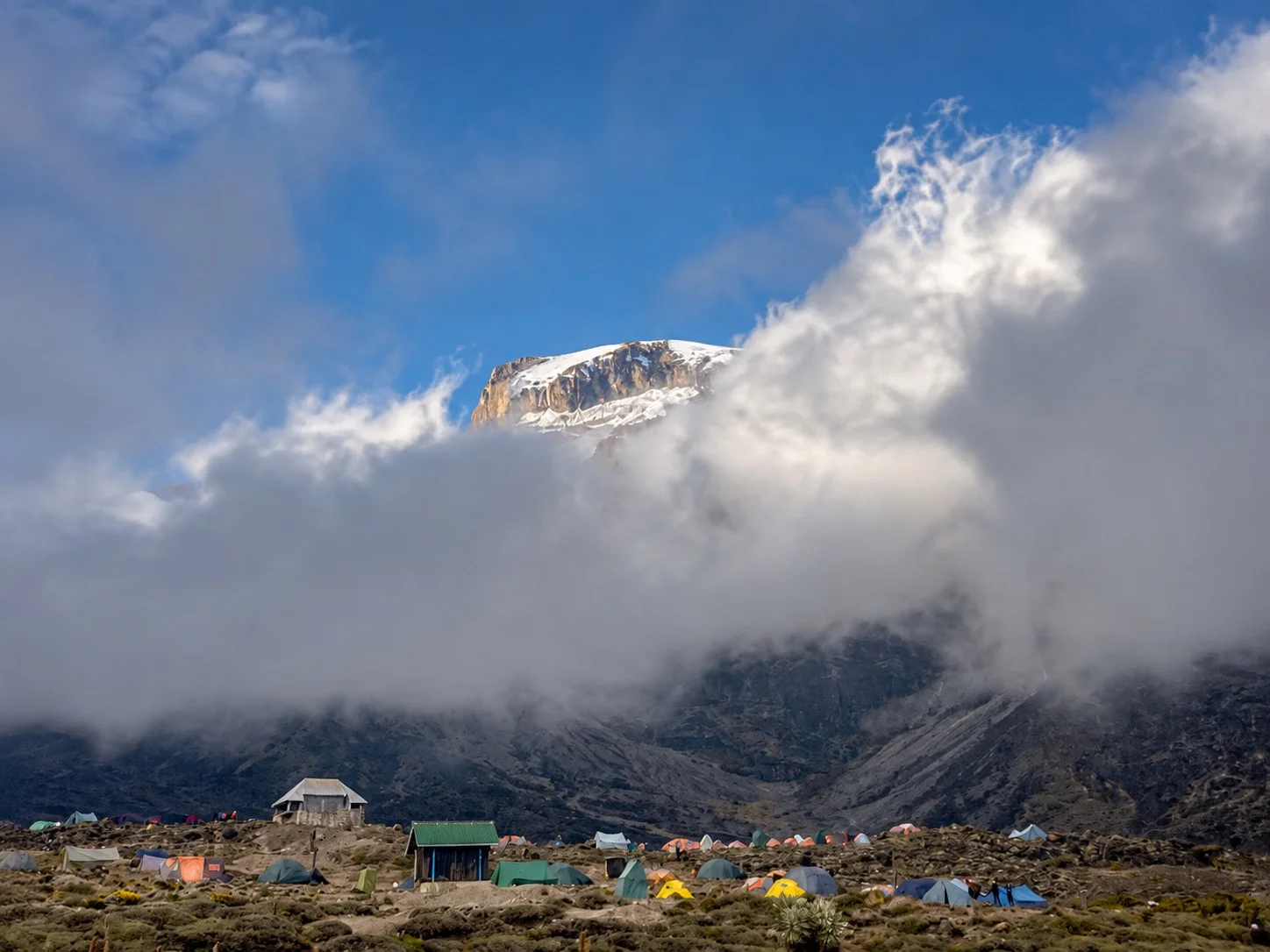 Mount Kilimanjaro’s snowcapped summit emerging through clouds above a colorful mountain camp