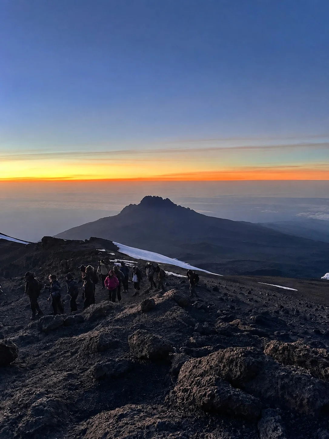 Climbers on Kilimanjaro beneath a vivid orange dawn and a distant mountain silhouette