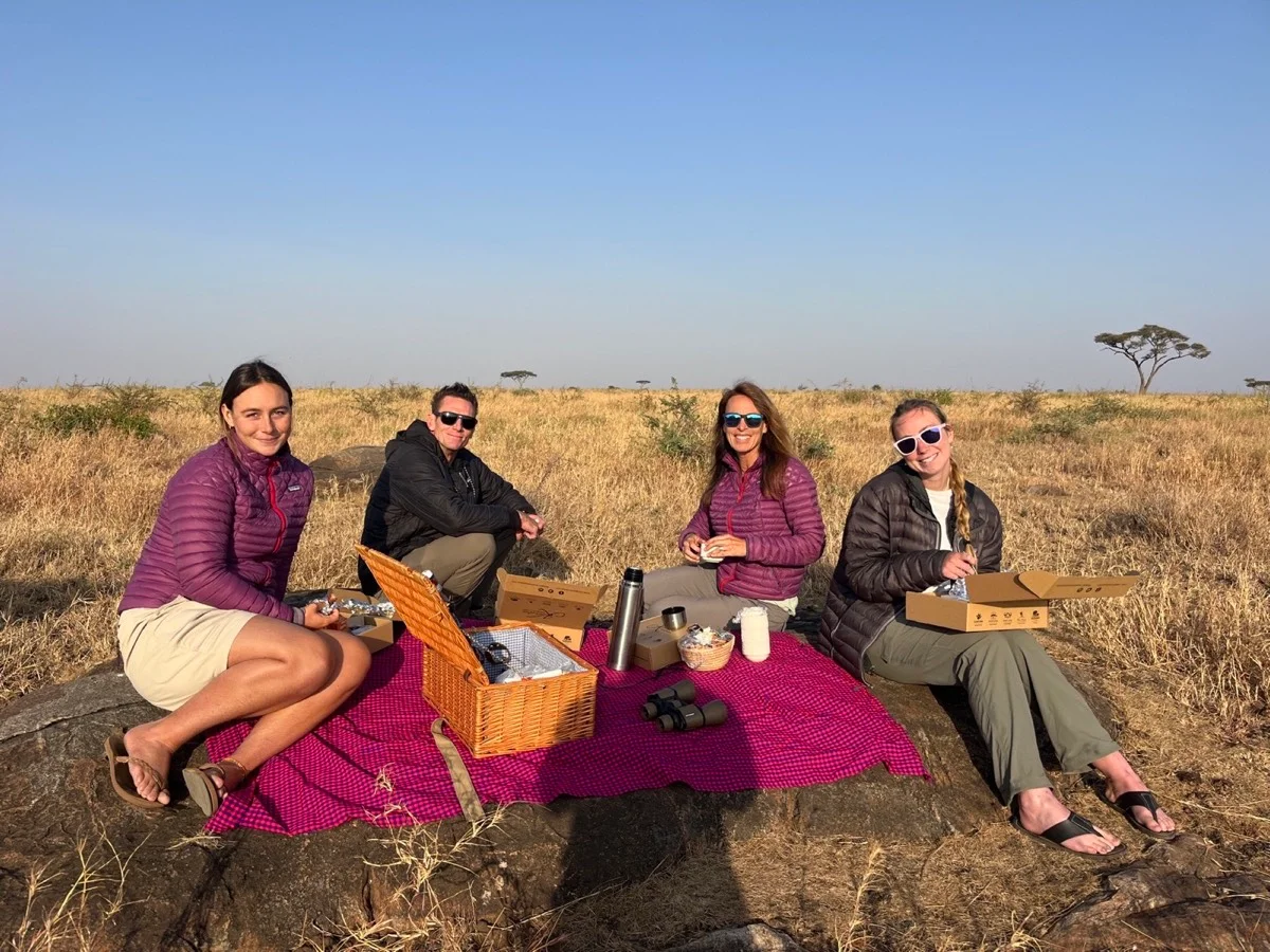 Four travelers sharing a picnic on a red and blue blanket in the open Serengeti landscape