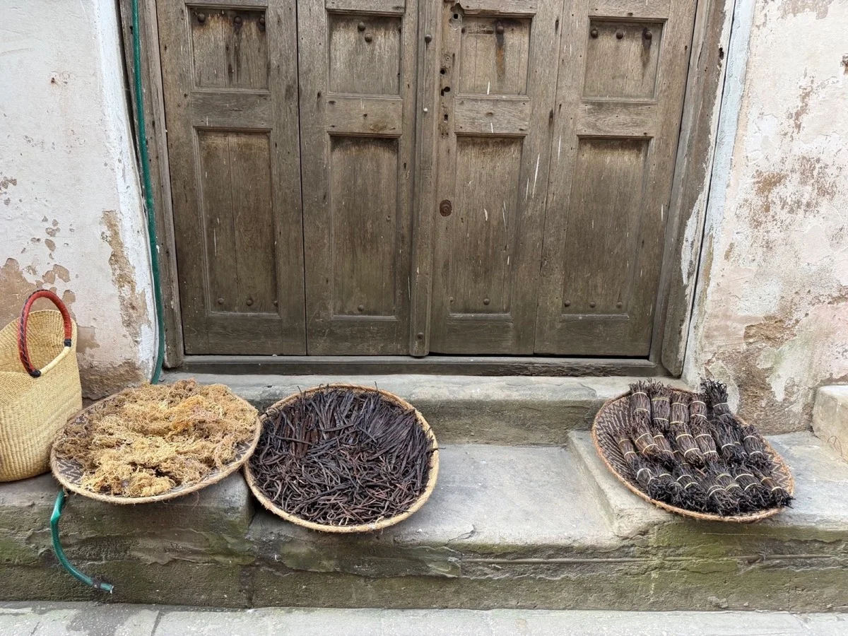 Woven trays of dried vanilla pods and local aromatics beside a weathered wooden door